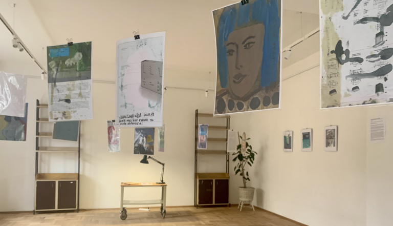 Galeria. Na pierwszym planie obrazki zawieszone na żyłkach. Na drugim planie stare biurko, na którym stoi zapalona metalowa lampka. Nad biurkiem wisi portrert mężczyzny w typie przewodniczącego partii. Wystawa Janusza Gałuszki - Kartoteka.