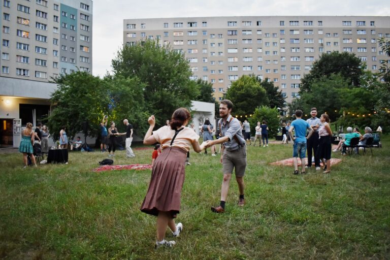 zdjęcie; mężczyzna i kobieta tańczący swinga. W tle bloki, zieleń oraz inni uczestnicy potańcówki