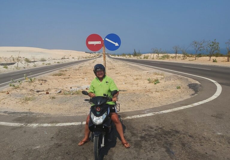 zdjęcie; uśmiechnięty mężczyzna w zielonej koszulce i czarnym kasku na czarnym motocyklu na zakręcie drogi na pustyni, za nim znaki drogowe