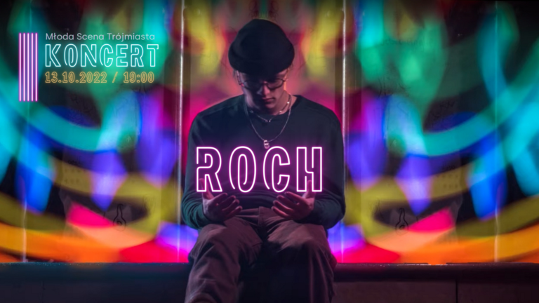 zdjęcie; młody mężczyzna ubrany na czarno siedzi na tle intensywnie kolorowej ściany, patrzy na swoje dłonie, nad którymi unosi się neon "Roch"