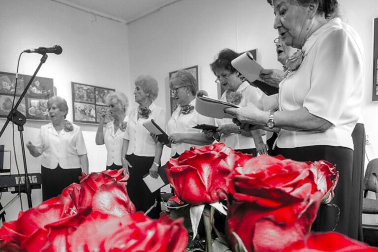 Czarnobiała fotografia śpiewających seniorek. Na dole czerwone róże.