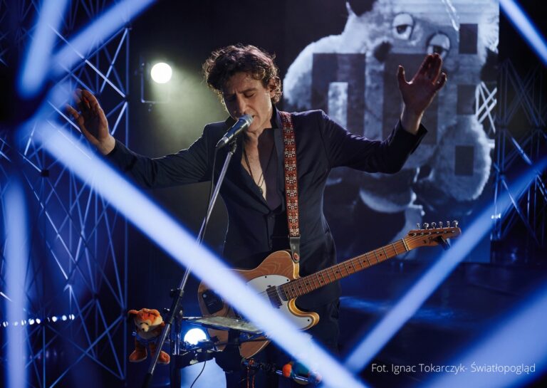 zdjęcie; mężczyzna z gitarą elektryczną stoi pośród scenografii, śpiewa do mikrofonu na statywie.
