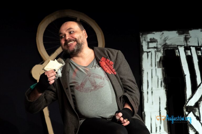 mężczyzna ubrany w t-shirt i marynarkę, na rękach ma rękawiczki z odkrytymi palcami, w jednej ręce trzyma kanapkę, jego paznokcie są pomalowane na czarno, patrzy na coś poza kadrem, ma zaciekawiony wzrok
