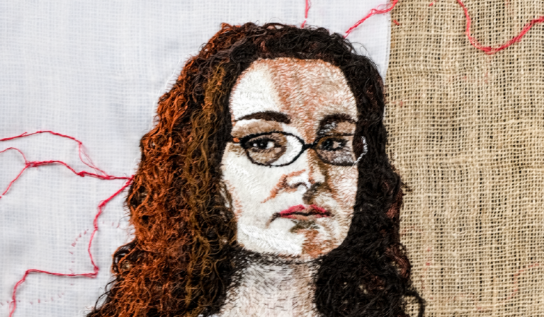 Artystyczny portret kobiety z ciemnymi, kręconymi włosami i okularami, wykonany techniką haftu. Tło składa się z białego płótna oraz fragmentu jutowego materiału, a czerwone nici biegną nieregularnie po powierzchni, nadając kompozycji ekspresyjny charakter.