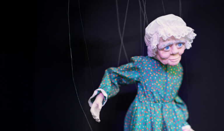 Marionetka przedstawiająca starszą kobietę w zielonej sukience w drobne kolorowe kropki. Laleczka ma jasną twarz, niebieskie oczy i biały czepek z koronką na głowie. Do postaci przymocowane są cienkie sznurki sterujące, widoczne na tle ciemnej, czarnej scenografii.