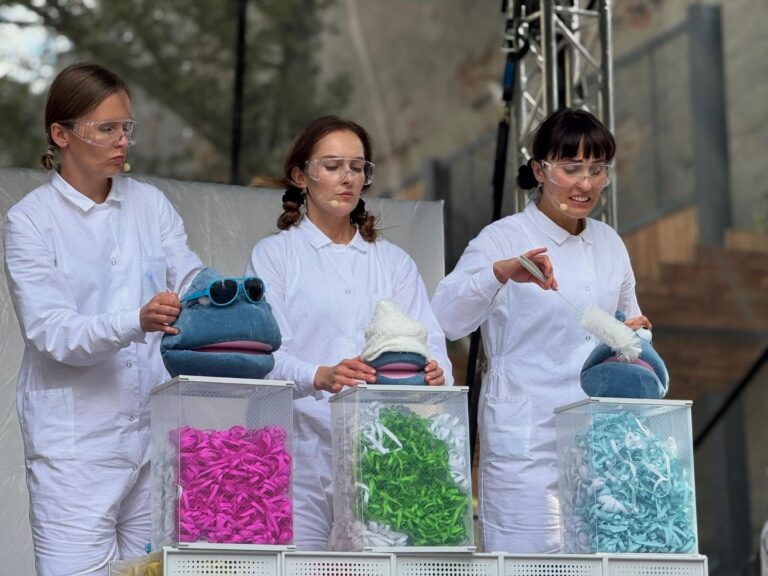Trzy kobiety stoją przy przezroczystych pojemnikach wypełnionych kolorowymi paskami papieru. Mają na sobie białe stroje i okulary ochronne. Trzymają niebieskie lalki i wykonują z nimi różne czynności, jakby prowadziły eksperyment lub pokaz.