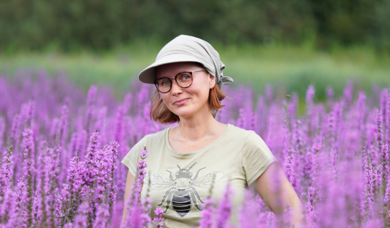 Zdjęcie: na łące pełnej lawendy stoi kobieta w okularach i chustce na głowie.