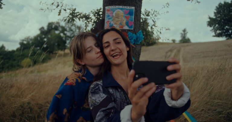 Para robi sobie selfie stojąc pod drzewem, do którego przypięty jest kolorowy obrazek. W tle pole z suchą trawą.