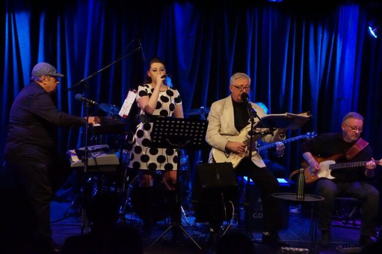 Zespół muzyczny podczas koncertu na scenie. Wokalistka śpiewa do mikrofonu, obok niej muzycy grają na instrumentach klawiszowych i gitarach. Scenę oświetla niebieskie światło, a w tle widać ciemną kurtynę oraz zestaw perkusyjny.