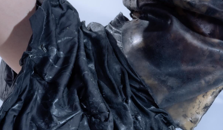 Zbliżenie na fragment ciemnego kostiumu teatralnego. Widoczne są liczne fałdy czarnej tkaniny oraz element przypominający rzeźbioną, ciemną maskę lub zbroję o gładkiej, błyszczącej powierzchni. Tło jest jasne i rozmyte.