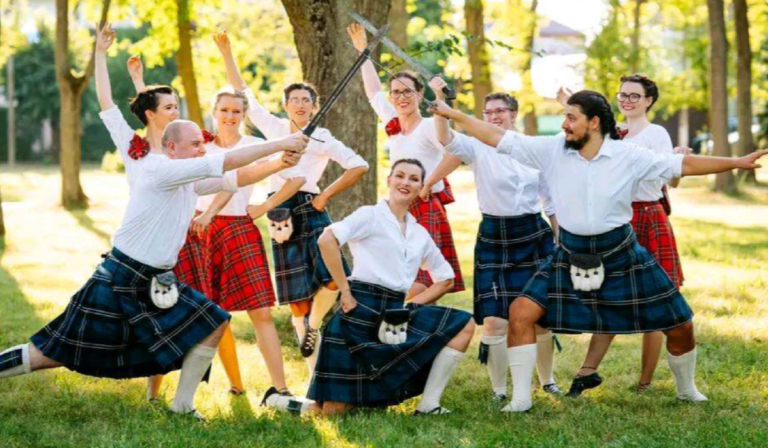 Grupa osób ubranych w białe koszule i szkockie kilty pozuje do zdjęcia wśród drzew.