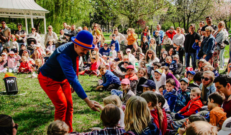 Artysta w niebieskim meloniku, granatowej koszuli i czerwonych spodniach występuje przed dużą grupą dzieci i dorosłych zgromadzonych na trawie w parku. Publiczność siedzi i stoi wokół niego, obserwując pokaz.